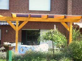 Holzpergola vor einem Backsteinhaus im Garten, mit Pflanzen im Vordergrund.