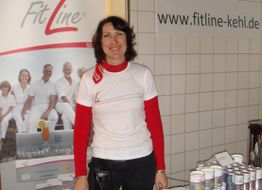 Frau in FitLine-Shirt steht an einem Stand mit Produkten. Banner und Webseite im Hintergrund.