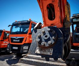 Drei orange-farbene Baufahrzeuge und ein Bagger mit "Powertilt"-Aufschrift bei sonnigem Wetter.