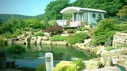 Garten mit Teich, Steinen und Pflanzen, kleines Haus im Hintergrund.