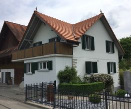 Weißes Haus mit rotem Dach, Balkon, Garten und Zaun an einem bewölkten Tag.
