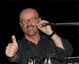 Ein Mann mit Brille zeigt Daumen hoch und spricht in ein Mikrofon, vor schwarzem Hintergrund.