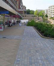 Fußweg mit Geschäften und Cafés links, Bäumen und Wohngebäuden im Hintergrund bei sonnigem Wetter.