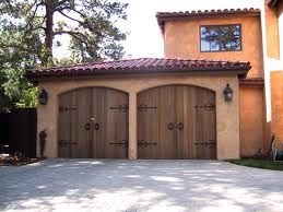 Two wooden garage doors on a stucco house with a tile roof and trees in the background.