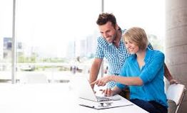 Duas pessoas sorrindo, olhando para um laptop em um escritório iluminado pelo sol.