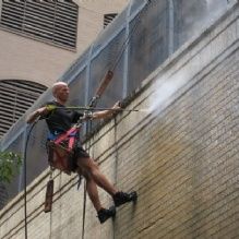 Man in safety harness power washing tall brick building.