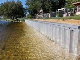 Metal seawall by a sandy shore with clear water, trees, and a fenced area in the background.