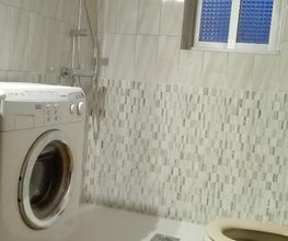 Baño con lavadora, inodoro y ducha. Azulejos blancos y ventana al fondo.