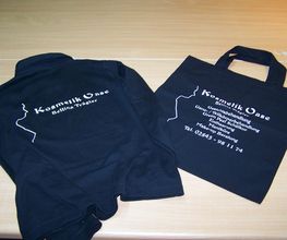 Schwarzes Hoodie und Tasche mit "Kosmetik Oase" Logo und Kontaktdetails auf Holzoberfläche.