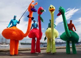 Bunte Figuren auf Stelzen als Vögel verkleidet vor einem Hafenhintergrund unter blauem Himmel.
