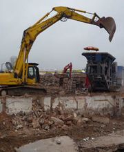 Bagger und Schuttschredder auf Baustelle, grauer Himmel, Ziegelreste im Vordergrund.