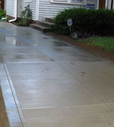 Wet concrete driveway next to a house with shrubs and grass along the edge.