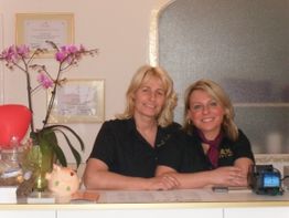 Zwei lächelnde Frauen hinter einem Empfangstresen mit Orchidee und Dekoration.