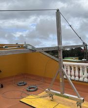 Grúa de metal en una terraza con cielo nublado y un pequeño asador al fondo.