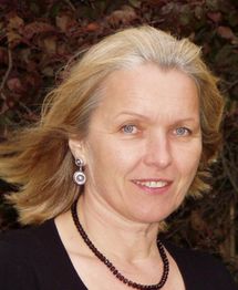 Eine Frau mit blonden Haaren, schwarzen Ohrringen und Kette lächelt in die Kamera vor Herbstlaub.