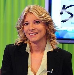 Eine lächelnde Frau mit welligem Haar in einem Anzug vor einem grünen Hintergrund und einem Bildschirm.