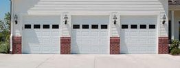Three white garage doors with brick accents and exterior lamps.