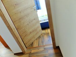 Geöffnete Holztür führt in ein helles Zimmer mit Holzboden und blauen Vorhängen.