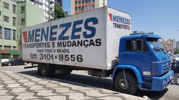 Caminhão azul da Menezes Transportes e Mudanças estacionado na rua em dia ensolarado.