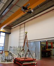 Industriehalle mit Brückenkran, Ketten und rotem Ladegut am Boden.