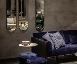 Elegantes Wohnzimmer mit blauem Sofa, Spiegeln und Hockern vor einer grauen Wand.