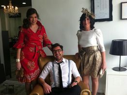 Persona sentada en sillón rodeada de dos mujeres de pie, sonrientes y vestidas elegantemente.