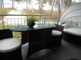 Balkon mit Tisch, zwei Stühlen und Pflanzen, Blick auf Sandweg und Bäume im Hintergrund.