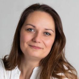 Eine Frau mit braunem Haar lächelt vor neutralem Hintergrund in die Kamera.