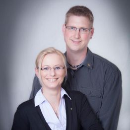 Zwei lächelnde Menschen mit Brillen, die vor einem neutralen Hintergrund posieren.