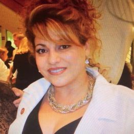 Woman smiling at a formal event, wearing a white coat and gold necklace.