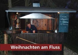 Ein Mann bedient einen Weihnachtsmarktstand am Fluss mit Currywurst-Angeboten.