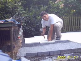 Mann legt Bodenfliesen im Freien, umgeben von Gartenzaun und Pflanzen.