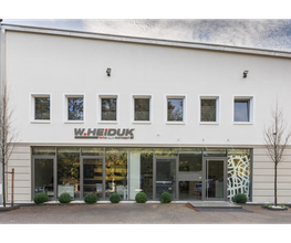 Moderne weiße Gebäudefassade mit sechs Fenstern und einem Firmenschild, umgeben von Bäumen.