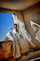 Eine Frau in weißem Kleid posiert in einem verlassenen Gebäude, blaue Himmel im Hintergrund.