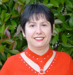 Donna con capelli corti neri e maglia rossa, davanti a piante verdi.