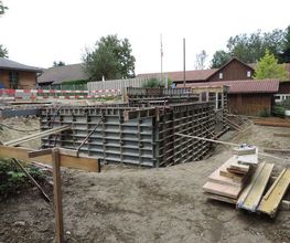 Baustelle mit Schalungen und Holzpaletten, umgeben von Gebäuden und Bäumen im Hintergrund.
