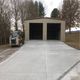 A two-door garage with a wide concrete driveway, a small vehicle parked on the gravel side.