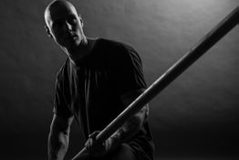 Mann in Schwarz-Weiß hält Holzstab, kraftvolle Pose, Licht und Schatten betonen die Szene.