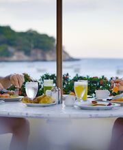 Pareja desayunando en una mesa con vista al mar; hay platos de fruta y bebidas.