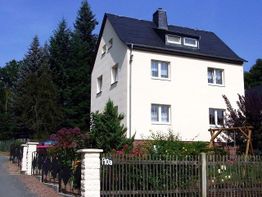 Zweistöckiges Haus mit Garten, Giebel und dunklem Dach, umgeben von hohen Bäumen und Zaun.