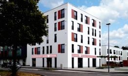Modernes, mehrstöckiges Gebäude mit weißen Wänden und roten Fensterläden an einer Straße.