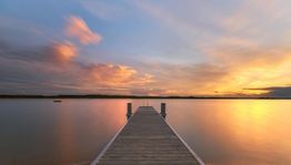 Steg am See bei Sonnenuntergang, leuchtend orangefarbener Himmel und ruhiges Wasser.