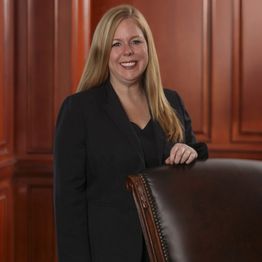 Woman in a black suit, standing and smiling near a brown leather chair, wood-paneled background.