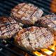 Four grilled burger patties sizzling on a barbecue with visible grill marks and flames.