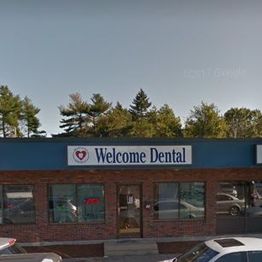 Red-brick building with "Welcome Dental" sign, surrounded by parked cars and trees in the background.