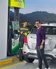 Hombre y empleada junto a auto en gasolinera, con montañas al fondo.
