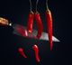 Knife slicing hanging red chilies against a dark background.