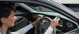 Driver using a phone while holding the steering wheel, seen inside a stationary car.