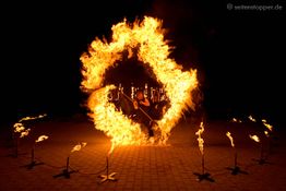 Feuershow mit Person in Flammenkreis, Nachtkulisse und mehreren Fackeln am Boden.