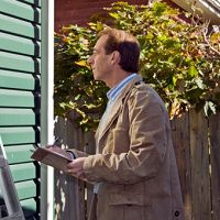 Man inspecting house exterior with a clipboard in hand, standing near green siding and a fence.